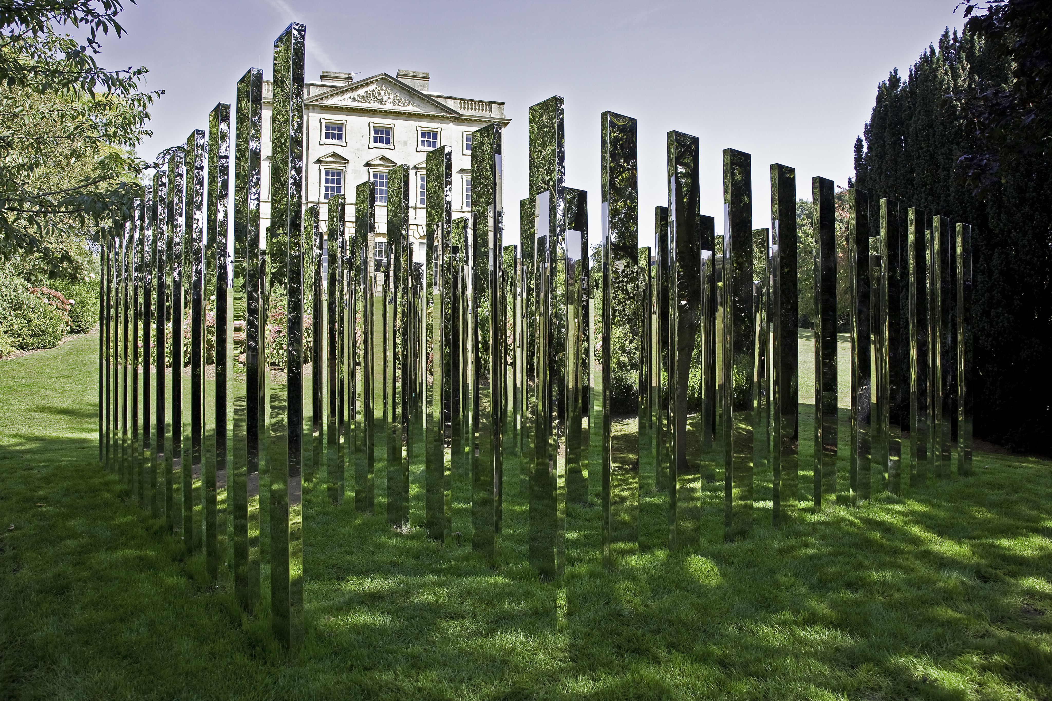 Follow Me (2019) by Jeppe Hein is shown on a sunny day in front of Royal Fort House. The artwork is a series of tall mirrored pillars, arranged in a rectangular fashion.
