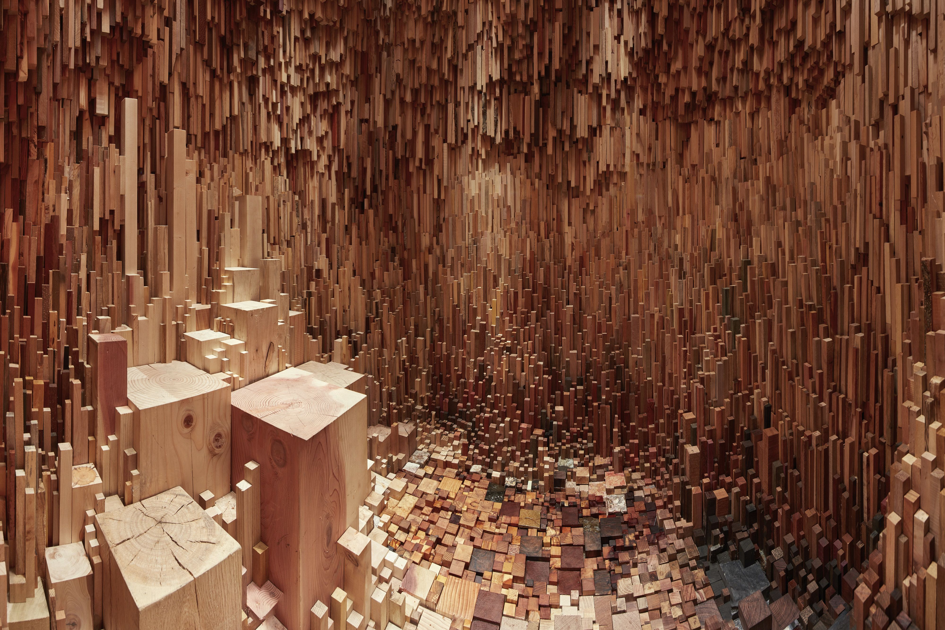 The inside of Hollow, an artwork created from wooden posts of varying sizes, heights and shades. Taller, wider blocks are on the left, with some darker, thinner blocks on the right. Sunlight is lighting many of the blocks.